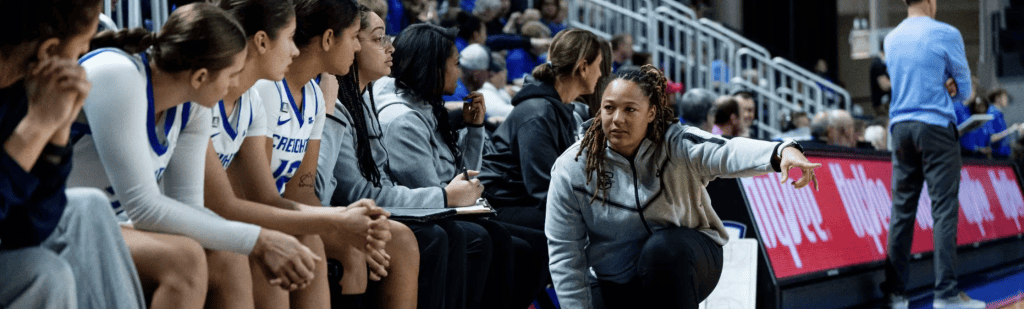 A basketball coach kneels beside seated players on the bench, giving instructions during a transformative journey in an indoor arena.